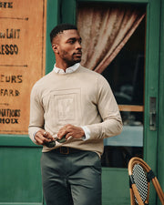 A man wearing the Psycho Bunny MENS OAK CITY CREW NECK - B6S274E200, a beige and lightweight sweater, over a white shirt stands outside a café. He holds sunglasses in one hand and gazes to the side, with a green door and part of a café chair visible in the background.
