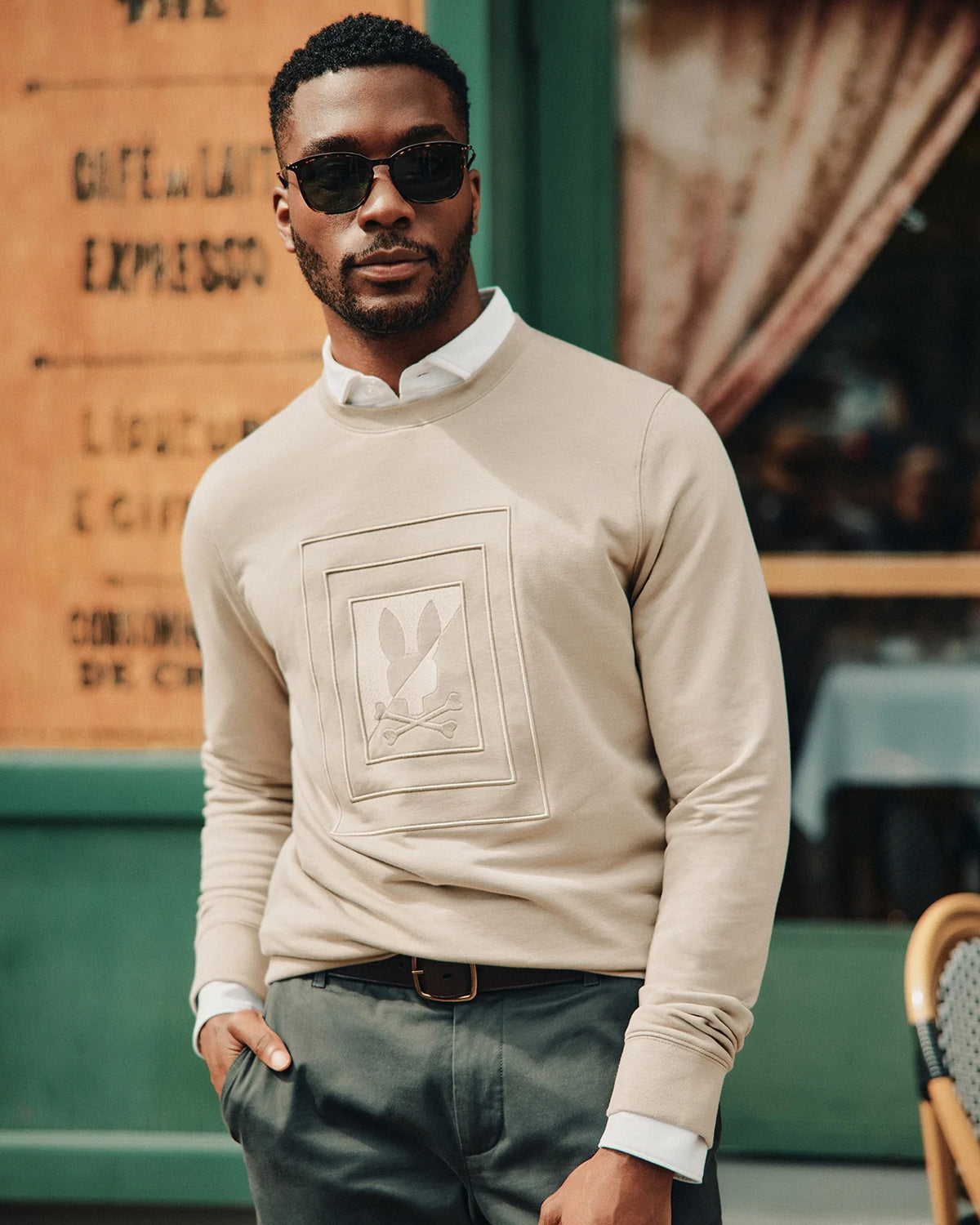 A man wearing Psycho Bunny's MENS OAK CITY CREW NECK - B6S274E200 and sunglasses stands confidently outside a cafe.