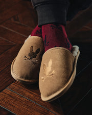 Close-up of a person wearing Psycho Bunny's MENS SUEDE SLIPPERS (B6A357E200) with genuine shearling lining and embroidered bunny designs. Paired with dark red socks featuring a leaf pattern, they rest on a polished wooden floor, exuding warmth and comfort.