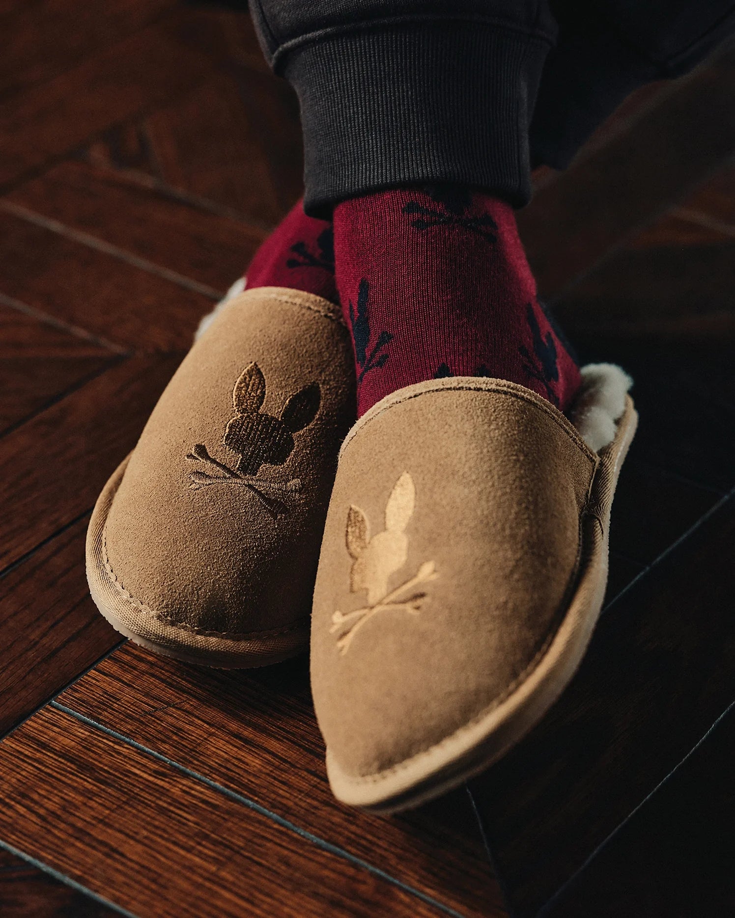 Close-up of a person wearing Psycho Bunny's MENS SUEDE SLIPPERS (B6A357E200) with genuine shearling lining and embroidered bunny designs. Paired with dark red socks featuring a leaf pattern, they rest on a polished wooden floor, exuding warmth and comfort.