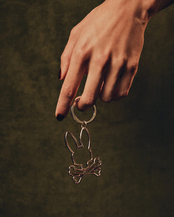 A hand with black-painted nails holds a Psycho Bunny OUTLINE BUNNY KEYCHAIN - B6A565E200, showcasing bunny ears and crossed bones, set against a dark green background.