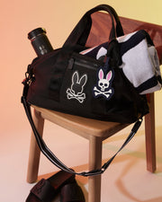 A black duffel bag with bunny skull and crossbones designs, sitting on a wooden chair. A black water bottle with a similar bunny motif is placed inside the bag, which also features a distinctive Psycho Bunny MENS LUGGAGE TAG - B6A387B200. A black and white striped towel is partially tucked into the bag, and black slides are on the ground.
