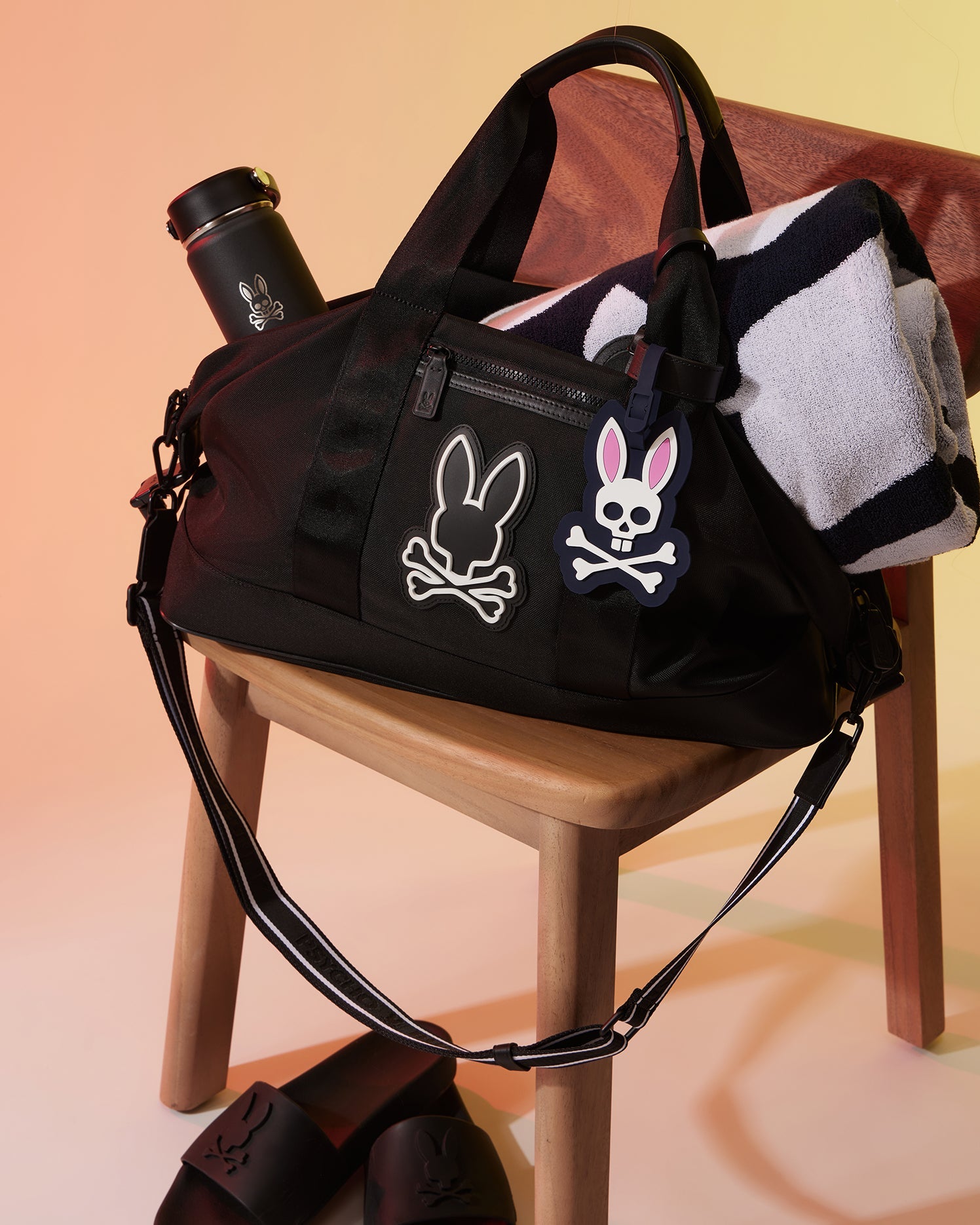 A black duffel bag with bunny skull and crossbones designs, sitting on a wooden chair. A black water bottle with a similar bunny motif is placed inside the bag, which also features a distinctive Psycho Bunny MENS LUGGAGE TAG - B6A387B200. A black and white striped towel is partially tucked into the bag, and black slides are on the ground.