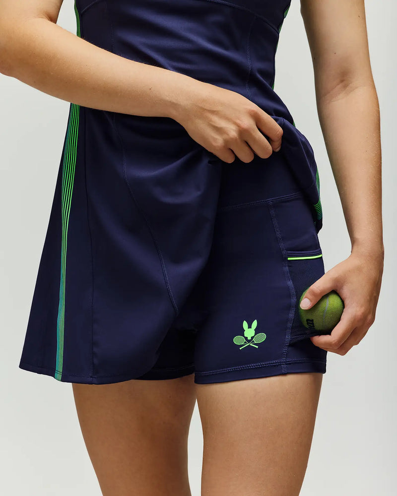 A person in the Psycho Bunny TENNIS BALL CREW WOMENS BIKER SHORT (WB0900001) lifts a navy blue athletic dress to reveal matching biker shorts with a green Bunny logo, holding a tennis ball in hand.