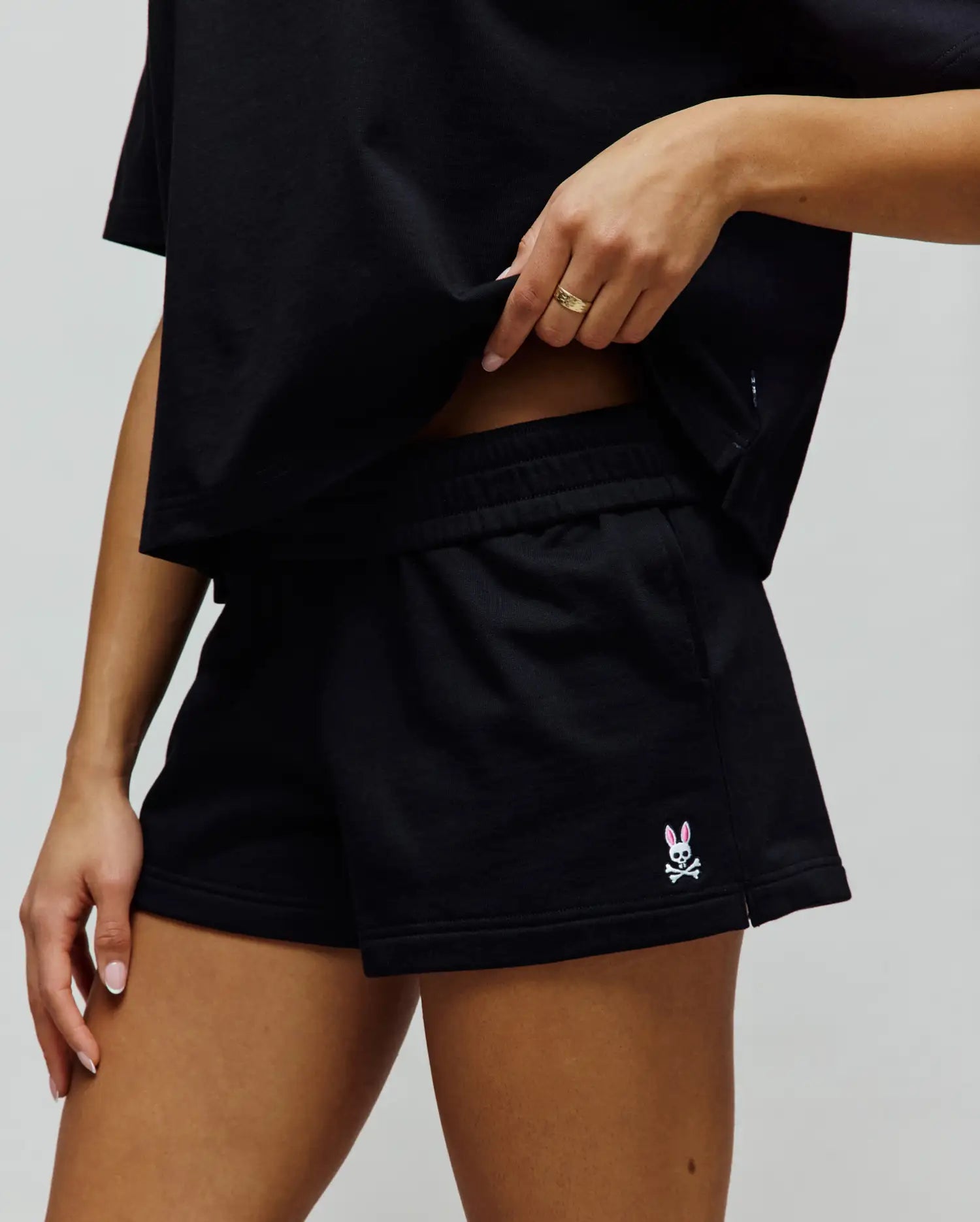 Womens Olivia Fleece Short, black relaxed-fit French terry shorts featuring a small embroidered Psycho Bunny skull logo with pink ears on the left leg hem.
