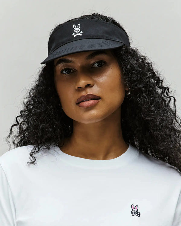 A woman with curly hair poses against a plain light background wearing the Psycho Bunny WOMEN'S SPORT VISOR - WO1500001, featuring a black design with a white bunny and crossbones logo, and a white shirt with a small pink bunny logo.