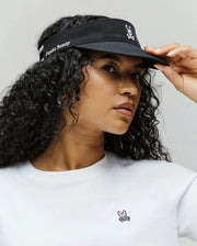 A woman with curly hair wears the Psycho Bunny WOMEN'S SPORT VISOR - WO1500001, black with an embroidered bunny logo and "Psycho Bunny" text, paired with a white tee featuring a small pink-and-black bunny logo on the chest.