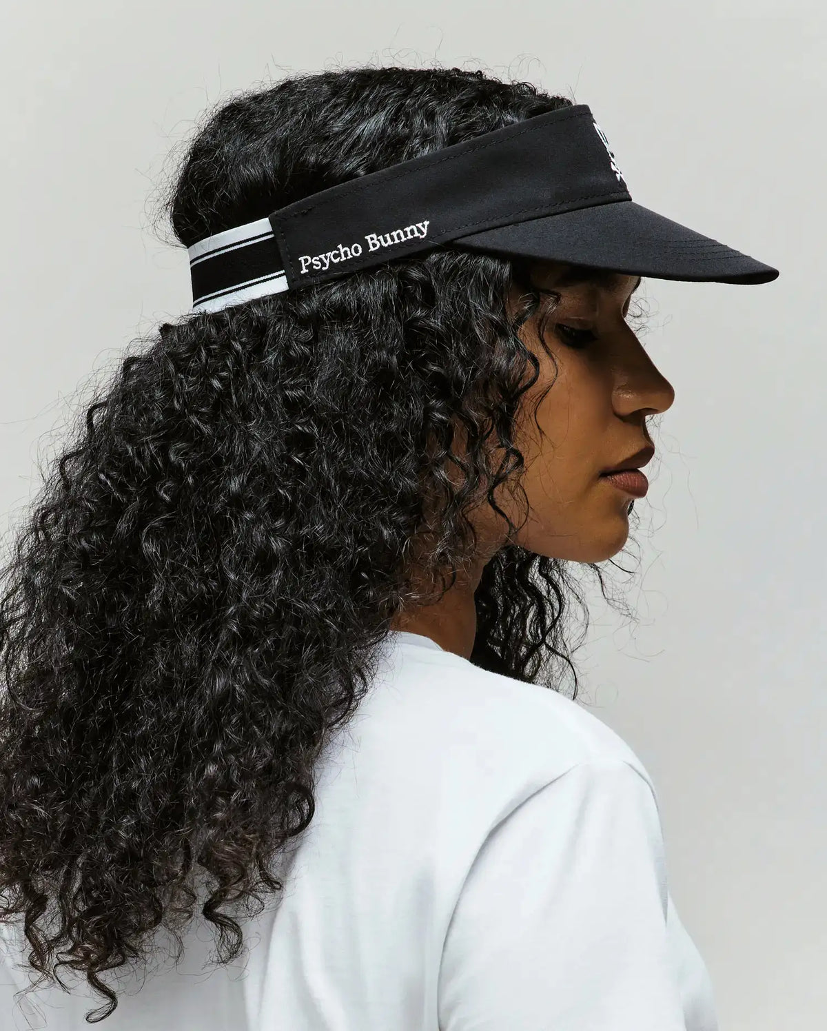A woman with long curly hair wears the Psycho Bunny WOMEN'S SPORT VISOR - WO1500001 in black, paired with a white shirt, shown in profile against a plain background.