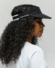 A woman with long curly hair wears the Psycho Bunny WOMEN'S SPORT VISOR - WO1500001 in black, paired with a white shirt, shown in profile against a plain background.