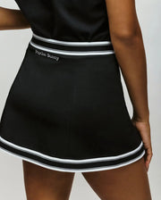 A person wears the Psycho Bunny WOMENS KNIT SKORT - WO1900001, a black skort with white stripes and "Psycho Bunny" embroidered on the waistband, standing with one hand resting at their side.