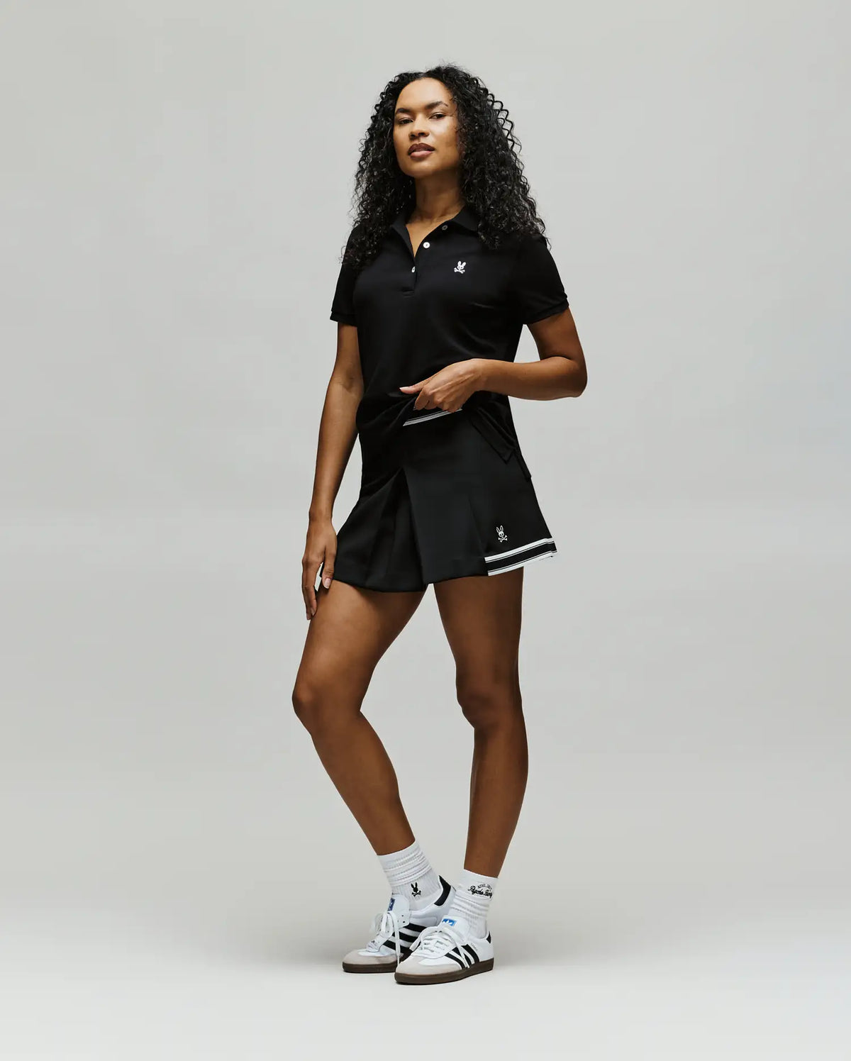 A confident woman with curly hair wears Psycho Bunny athletic wear: a black polo, WOMENS KNIT SKORT - WO1900001, white socks, and white sneakers, posing against a plain light background.