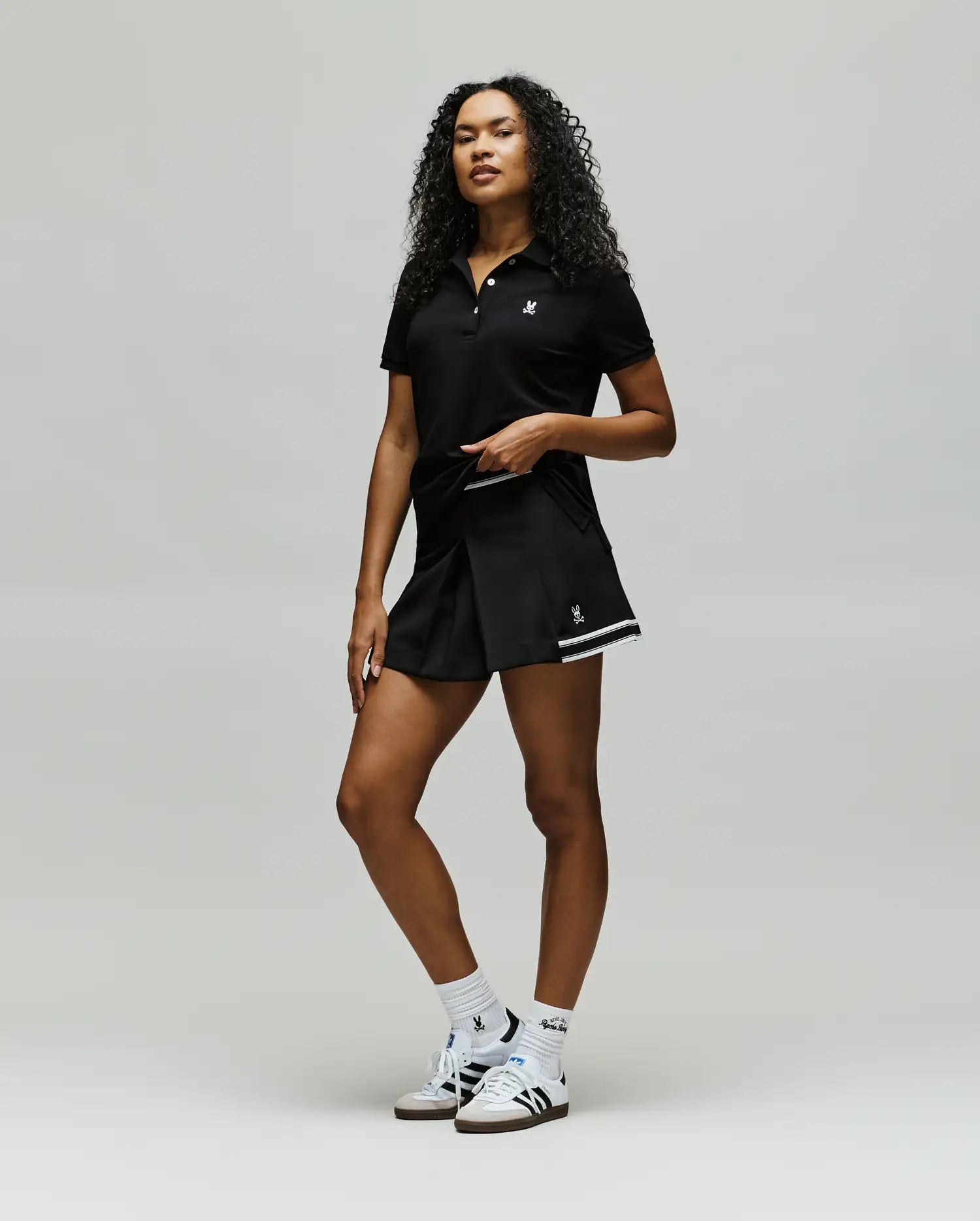A confident woman with curly hair wears Psycho Bunny athletic wear: a black polo, WOMENS KNIT SKORT - WO1900001, white socks, and white sneakers, posing against a plain light background.