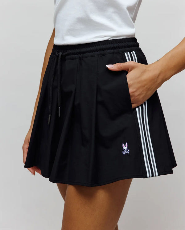 Womens Cecilia Pleated Skort in black, featuring a relaxed fit, smooth woven fabric, elastic drawstring waist, three white side stripes, and a small embroidered pink Psycho Bunny skull logo on the lower left.