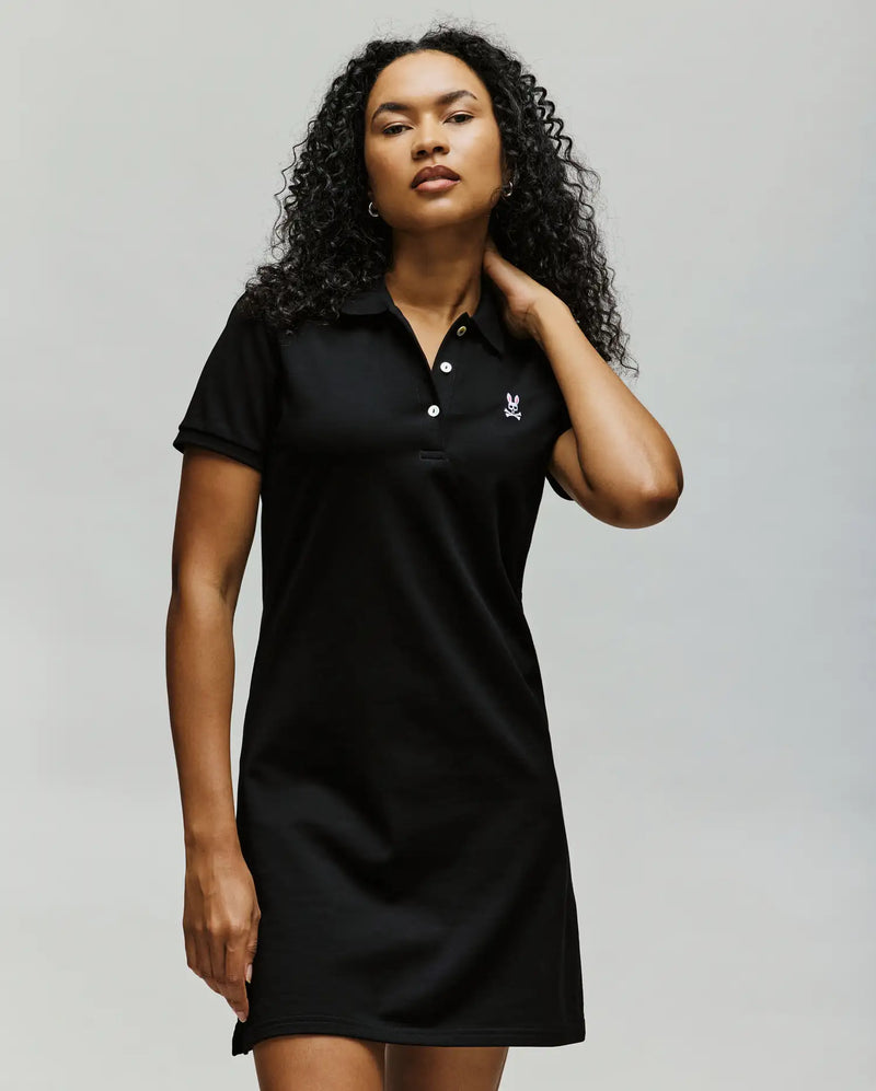 A woman with curly hair models Psycho Bunny’s WOMENS POLO DRESS - WO2000003, a short-sleeved black dress featuring a small embroidered Bunny logo on the chest, standing confidently against a light background.