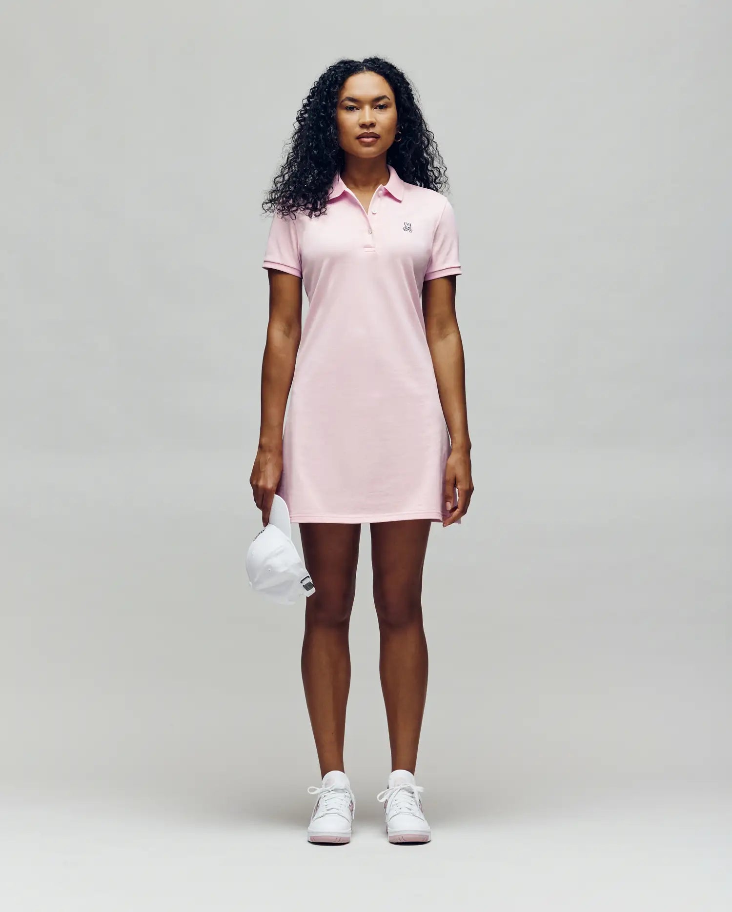 A person faces forward in a Psycho Bunny WOMENS POLO DRESS - WO2000003, a light pink polo dress with a feminine silhouette, styled with white sneakers and holding a white baseball cap against a plain light gray background.