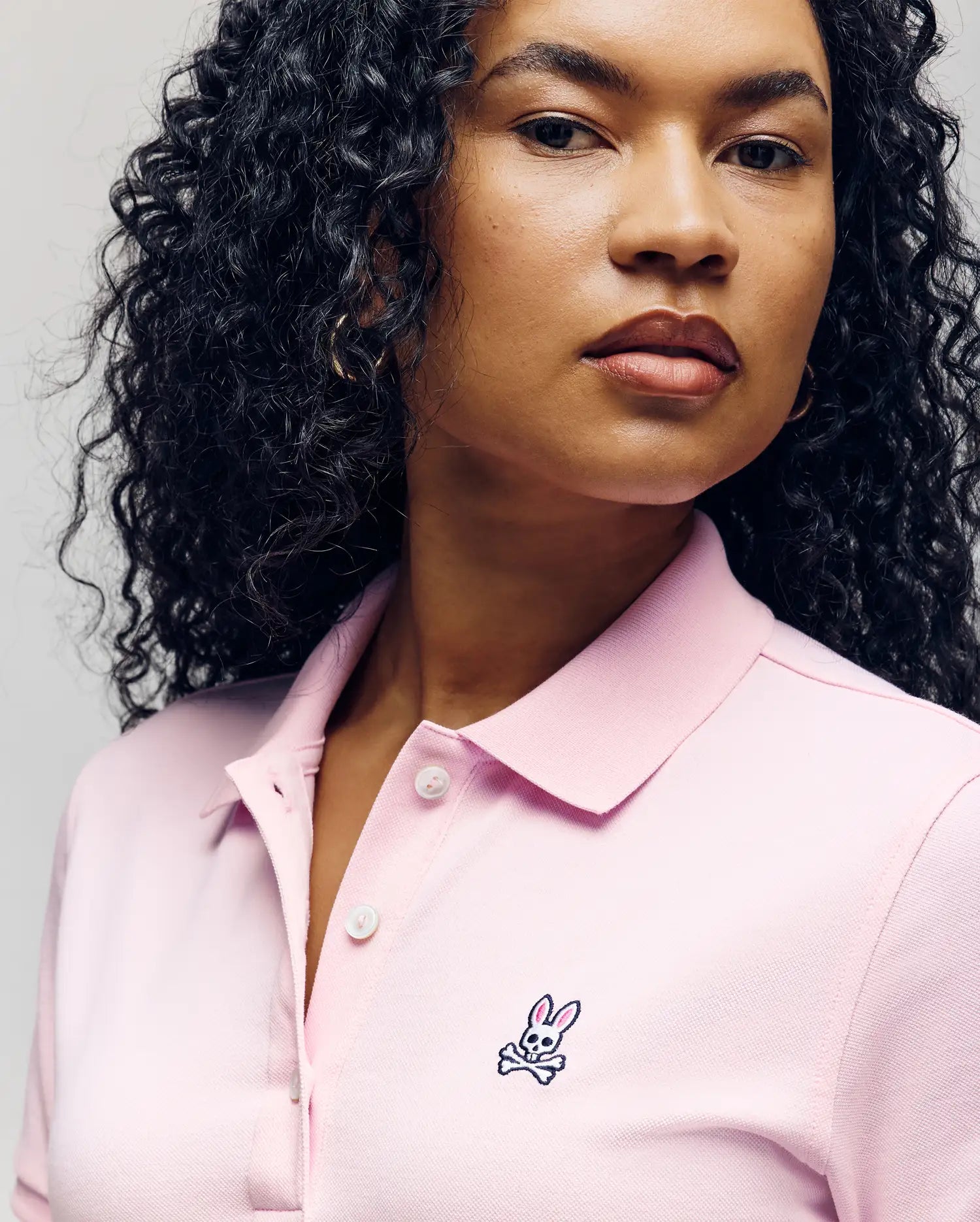 A woman with curly hair wears the Psycho Bunny WOMENS POLO DRESS - WO2000003 in light pink, featuring a feminine silhouette, Mother-of-Pearl buttons, and an embroidered bunny and crossbones logo on the chest.