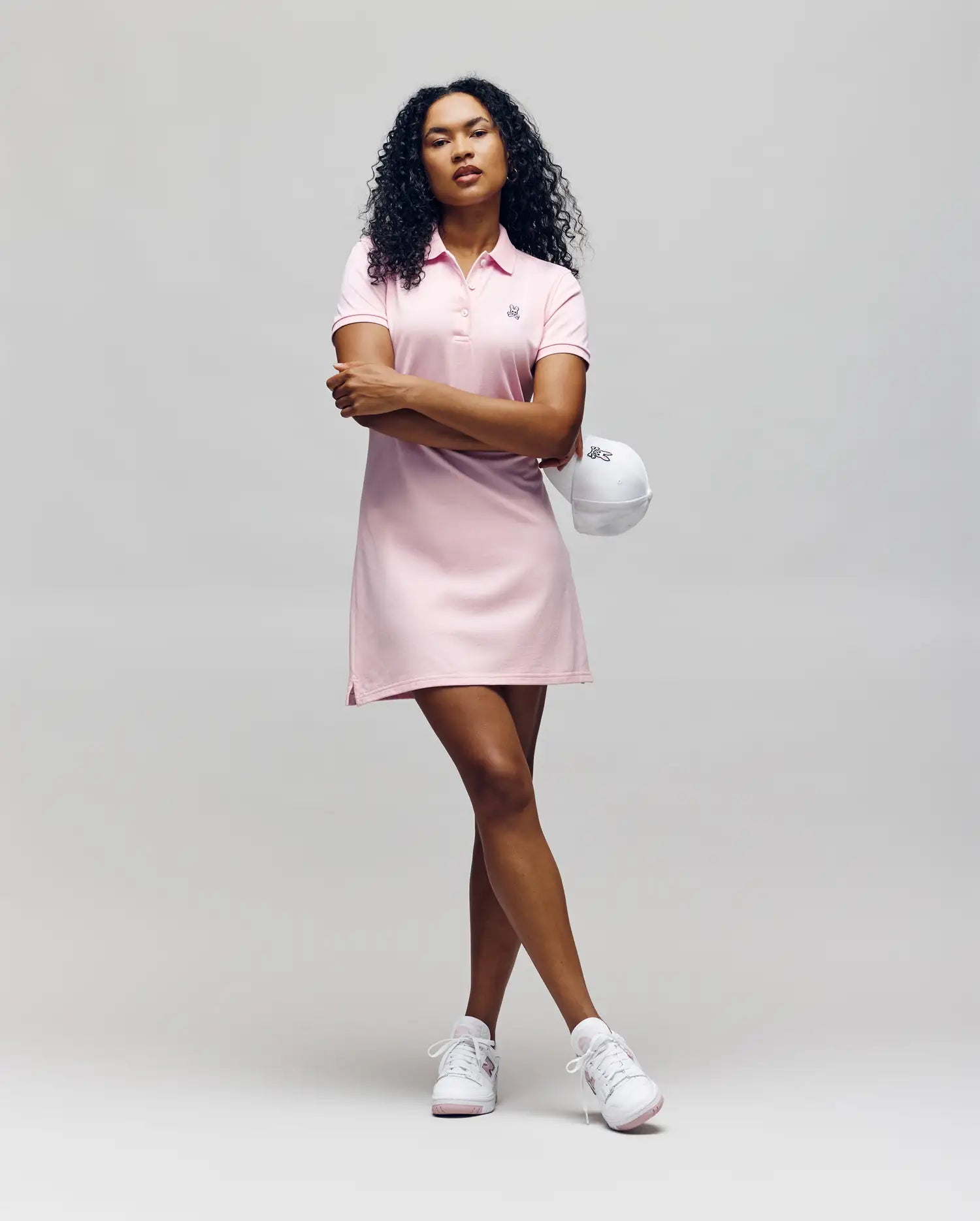 A confident person with curly hair wears the Psycho Bunny WOMENS POLO DRESS - WO2000003 in light pink, holding a white cap and pairing the look with classic white sneakers against a plain background.