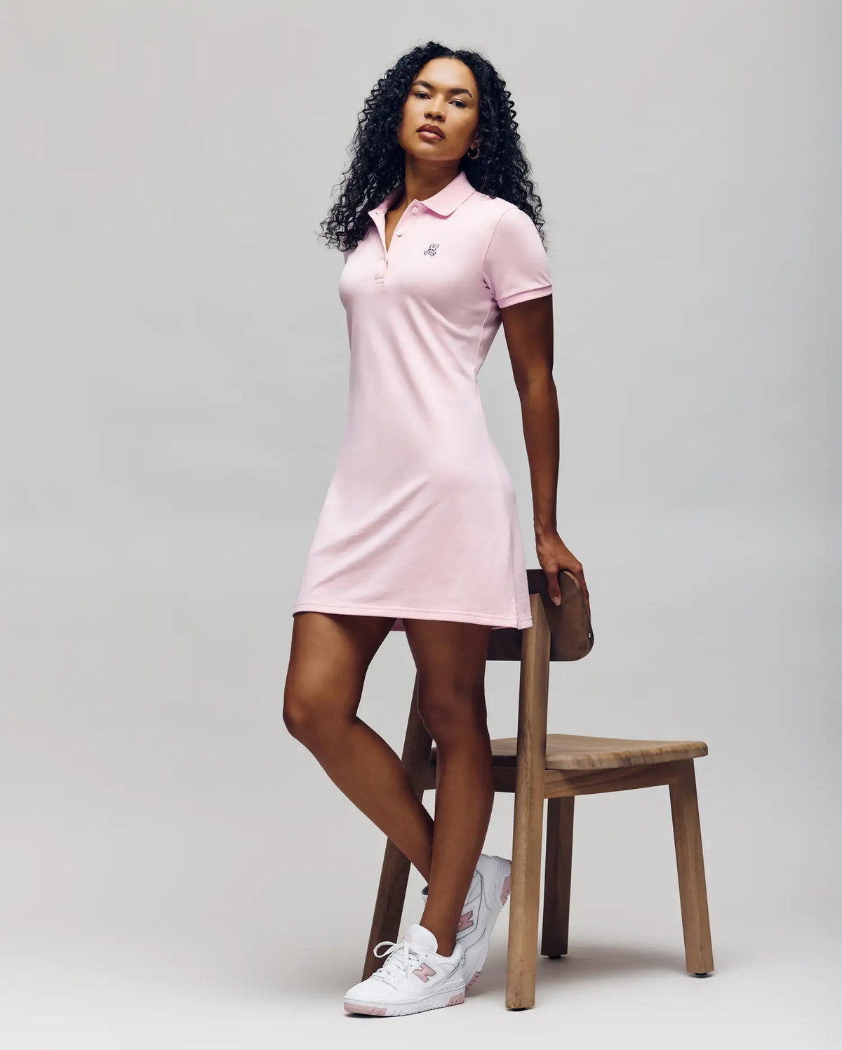 A woman with curly hair poses by a wooden chair, wearing the Psycho Bunny WOMENS POLO DRESS - WO2000003 in light pink with Mother-of-Pearl buttons and white sneakers, set against a plain light background.