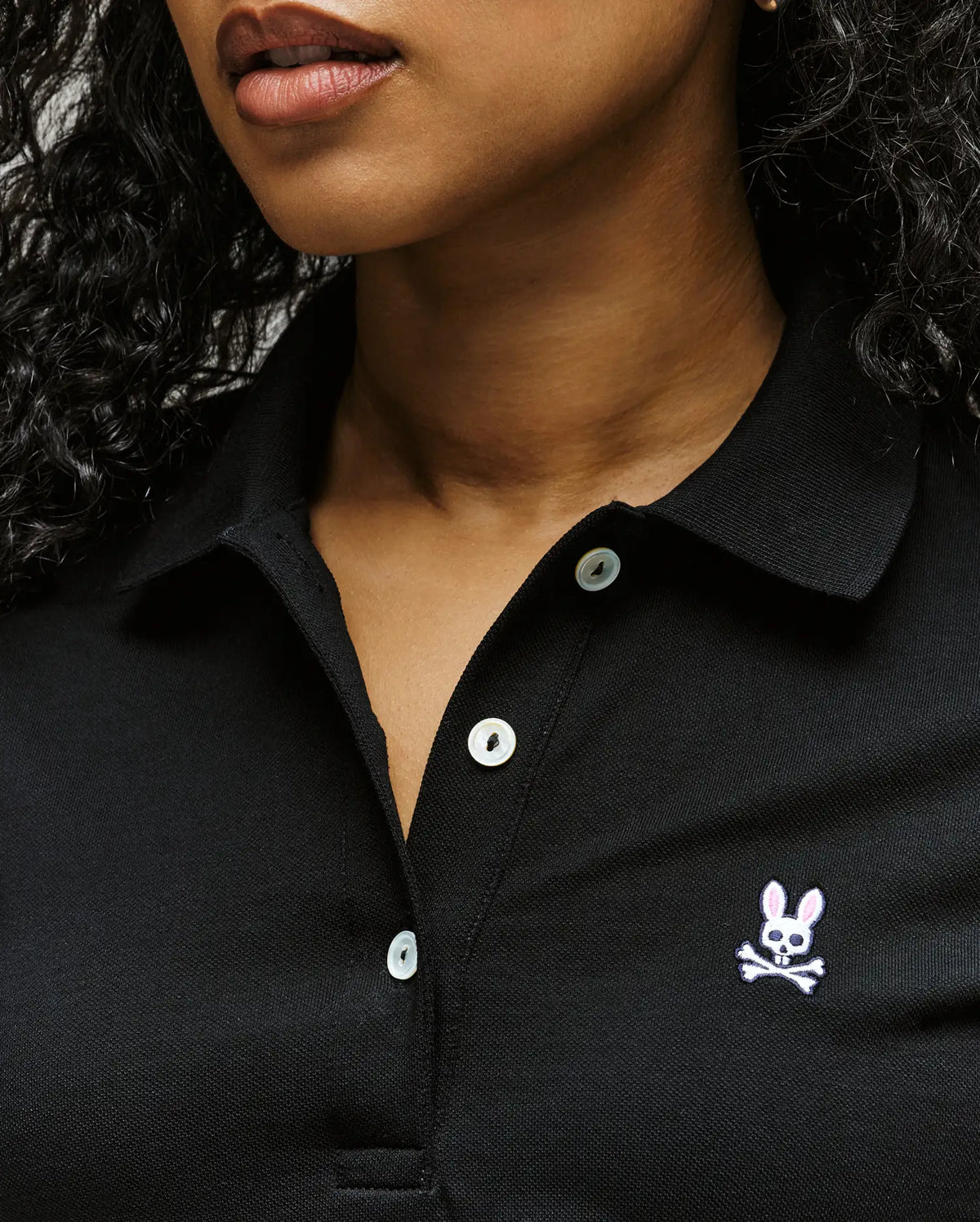 A close-up of a woman in the Psycho Bunny WOMENS PIQUE POLO - WT0100003, featuring white buttons and a 4k-stitch embroidered Bunny skull and crossbones logo; only her lips and lower face are visible.