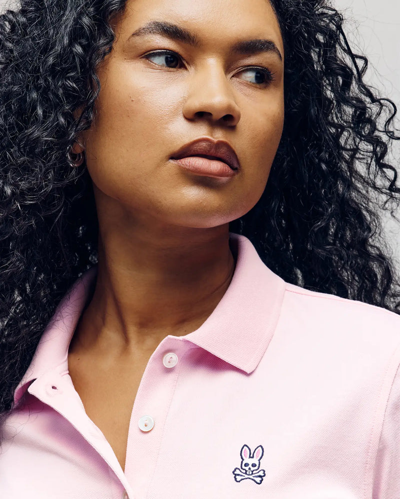 A woman with curly hair looks to the side in the Psycho Bunny WOMENS PIQUE POLO - WT0100003, a light pink shirt with an embroidered Bunny and crossed bones logo, offering a flattering fit and effortless style.