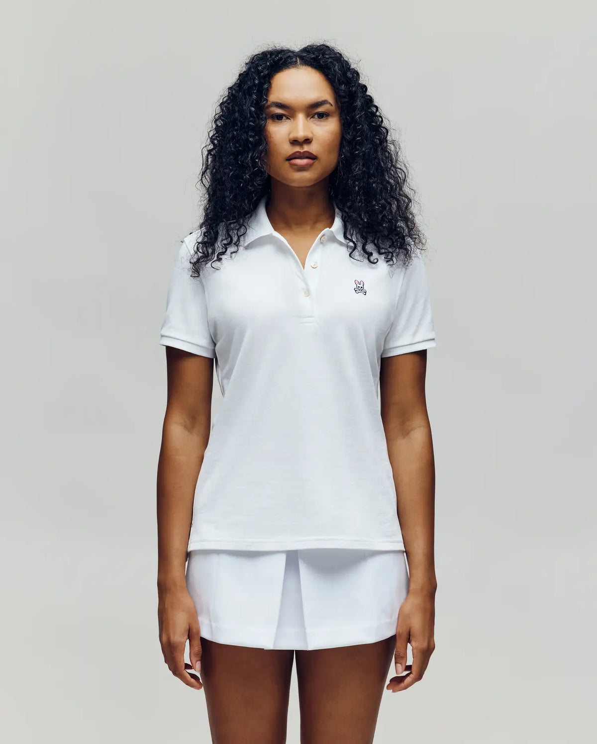 A woman with curly hair stands facing forward, wearing the Psycho Bunny WOMENS PIQUE POLO - WT0100003 in white and a white skirt against a light background.