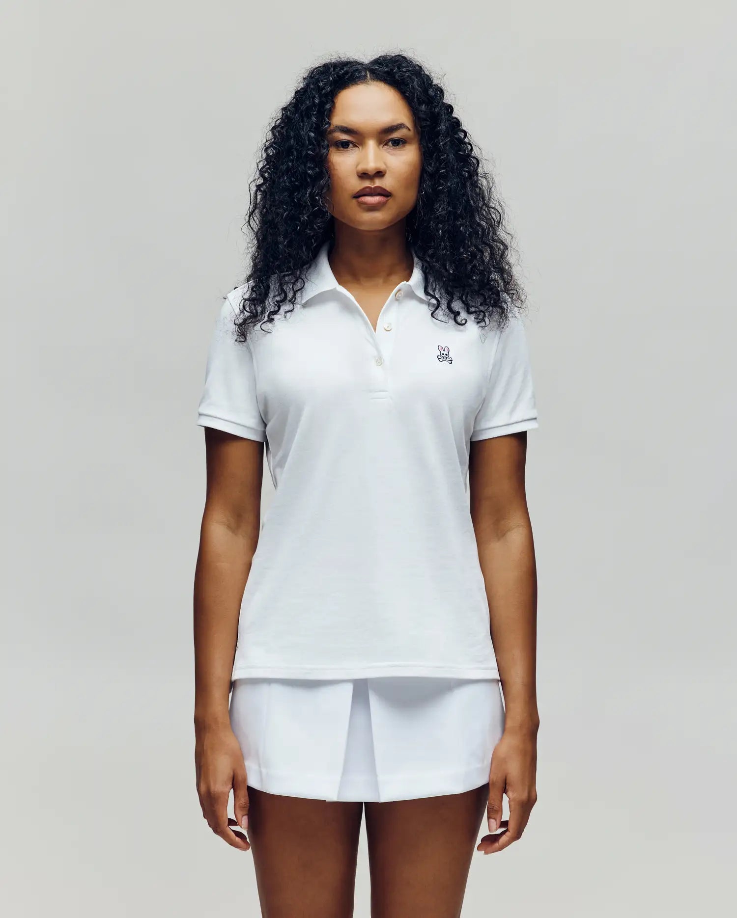 A woman with curly hair stands facing forward, wearing the Psycho Bunny WOMENS PIQUE POLO - WT0100003 in white and a white skirt against a light background.