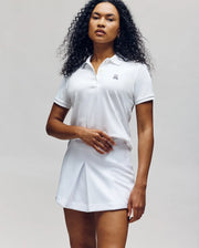 A person with long curly hair wears the Psycho Bunny WOMENS PIQUE POLO - WT0100003 in white and a white pleated skirt, standing against a plain light gray background.