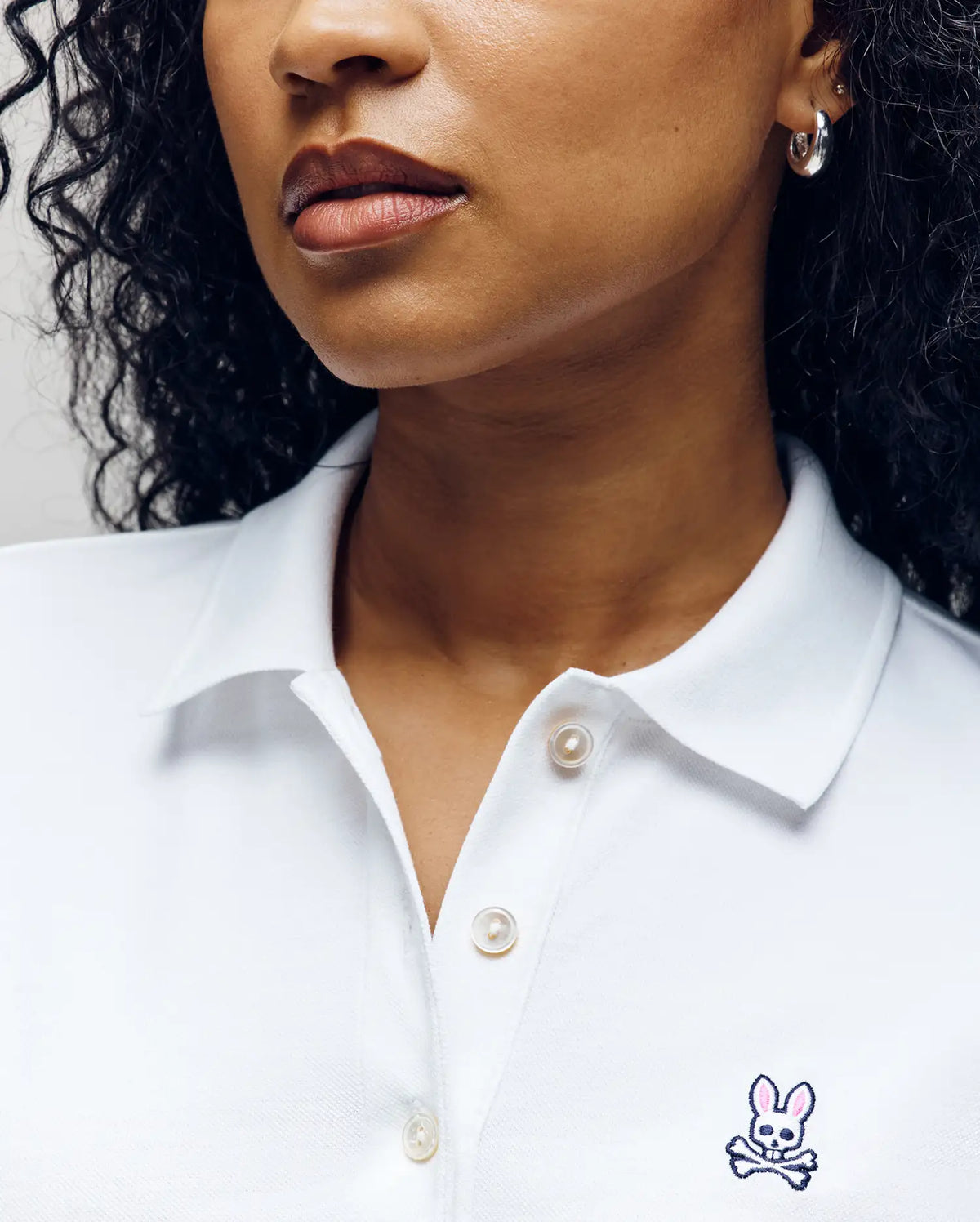 A woman with curly hair models the Psycho Bunny WOMENS PIQUE POLO - WT0100003 in white, showing only her lower face and upper torso. The shirt features a small embroidered Bunny with crossbones on the chest.