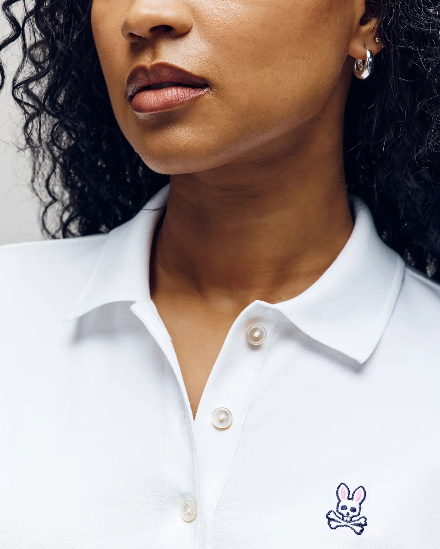 A woman with curly hair models the Psycho Bunny WOMENS PIQUE POLO - WT0100003 in white, showing only her lower face and upper torso. The shirt features a small embroidered Bunny with crossbones on the chest.