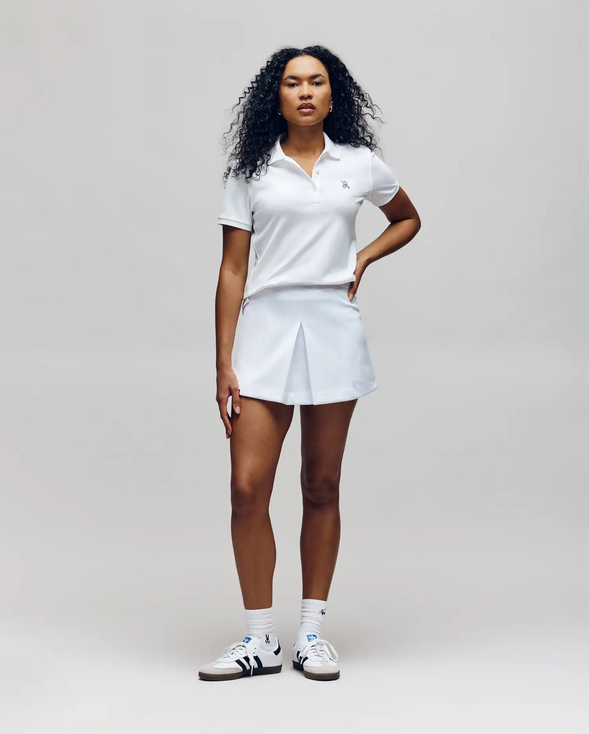 A woman with curly hair, facing forward, wears the Psycho Bunny WOMENS PIQUE POLO - WT0100003, paired with a white pleated skirt, white socks, and white sneakers with dark accents against a plain light background.