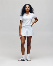 A woman with curly hair, facing forward, wears the Psycho Bunny WOMENS PIQUE POLO - WT0100003, paired with a white pleated skirt, white socks, and white sneakers with dark accents against a plain light background.
