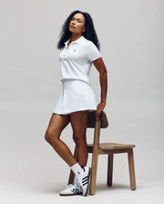 A woman in the Psycho Bunny WOMENS PIQUE POLO (WT0100003) and a tennis skirt leans against a wooden chair. She has curly hair, wears white socks and Adidas sneakers with black stripes, on a neutral background.