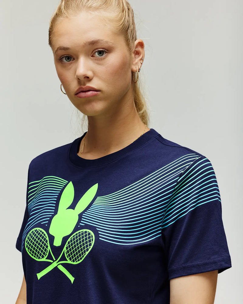 A young woman with blonde hair in a ponytail wears the Psycho Bunny TENNIS BALL CREW WOMENS TEE, featuring a neon green bunny head, crossed tennis rackets, and curved blue and green lines across the chest.