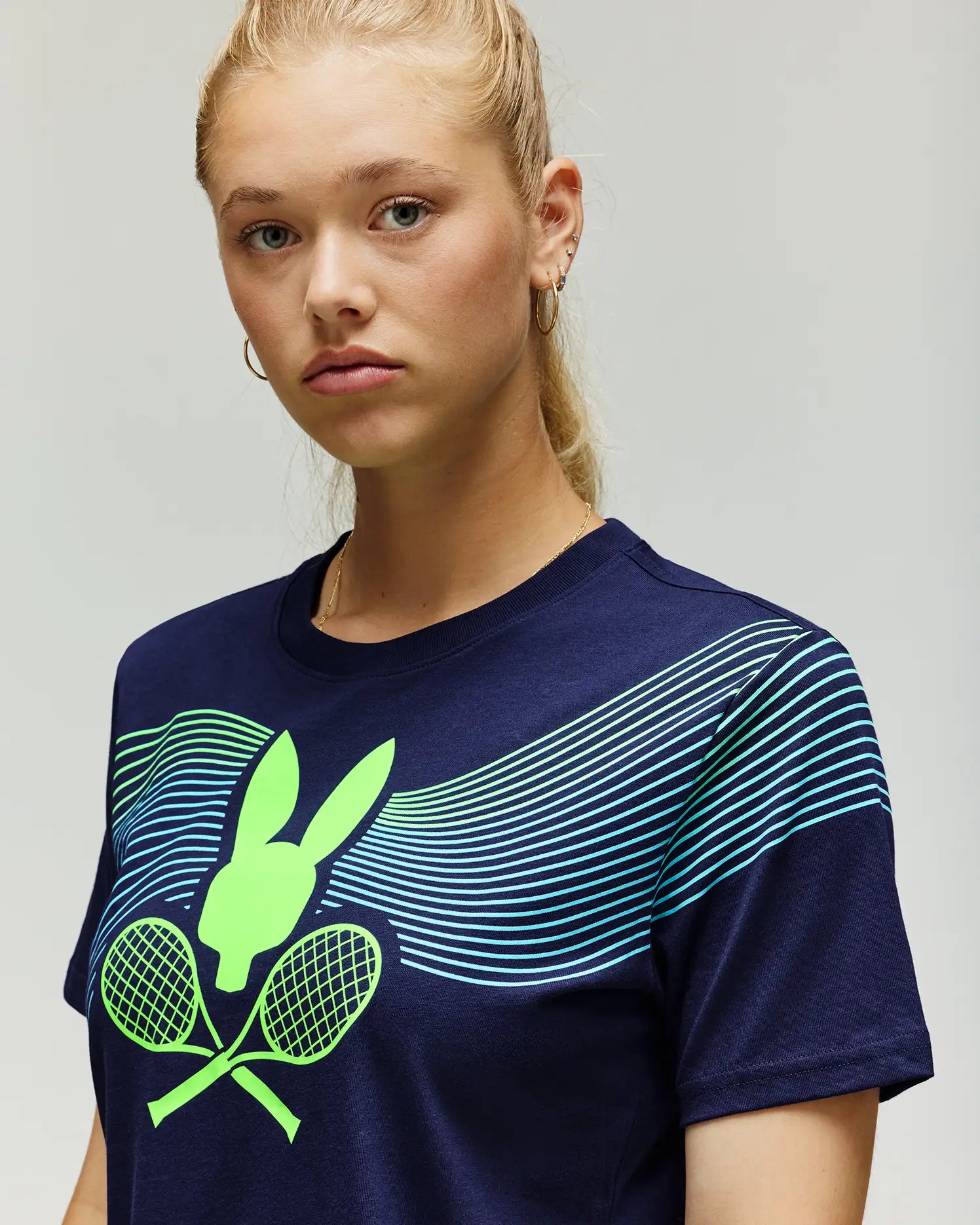 A young woman with blonde hair in a ponytail wears the Psycho Bunny TENNIS BALL CREW WOMENS TEE FRENCH (WT0200004), a navy Pima cotton shirt with a neon green Tennis Bunny graphic, crossed rackets, and blue-green chest lines.