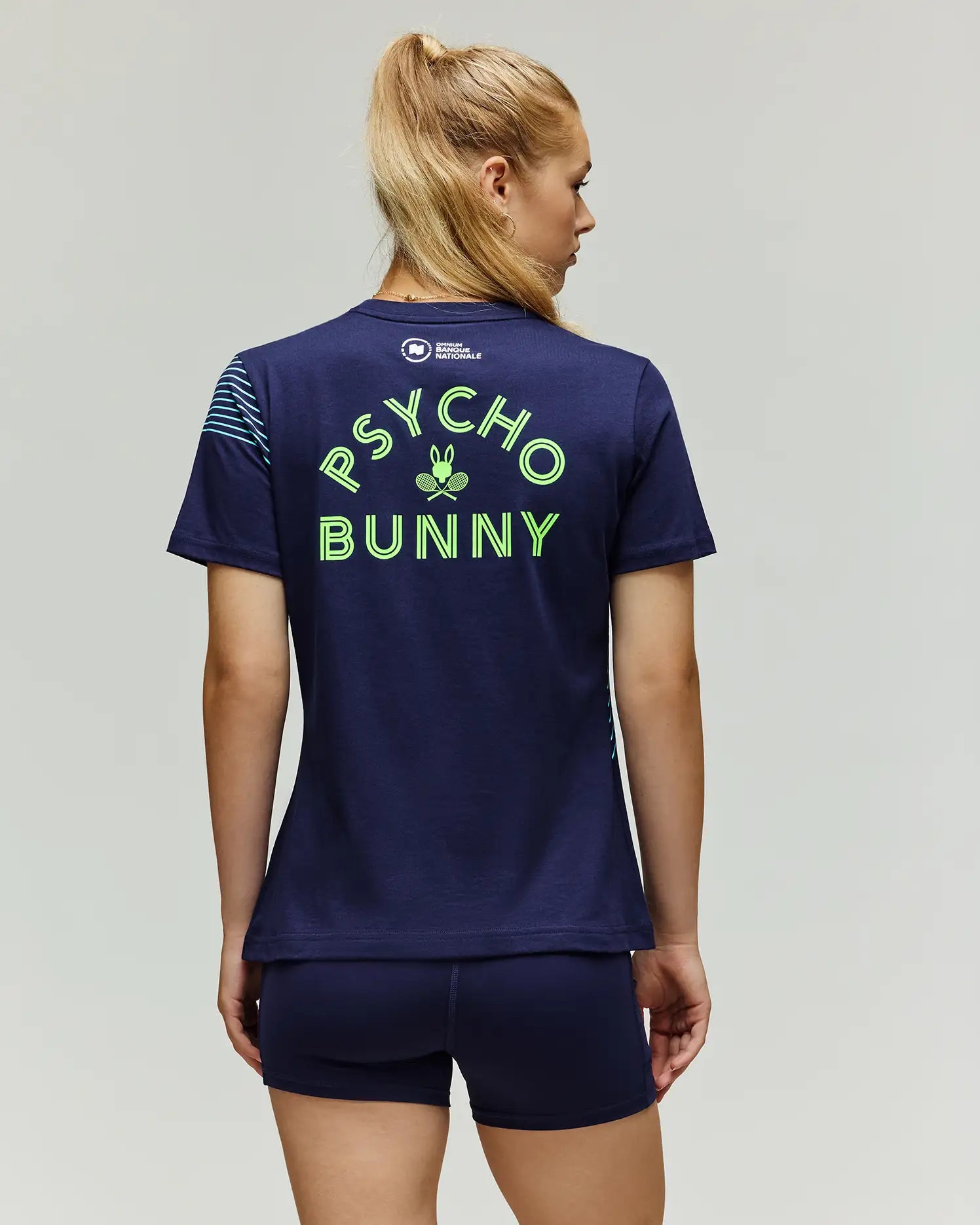 A woman with blonde hair in a ponytail is seen from behind wearing the Psycho Bunny TENNIS BALL CREW WOMENS TEE FRENCH (WT0200004), a navy Pima cotton tee with a neon green Tennis Bunny graphic on the back, paired with navy shorts.
