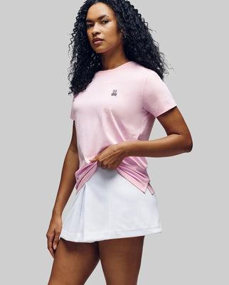 A woman with curly hair models the Psycho Bunny WOMENS CLASSIC TEE - WT0200005 in light pink, made from 100% Pima cotton, featuring a 4k-stitch embroidered Bunny. She pairs it with a white pleated skirt against a plain gray background.