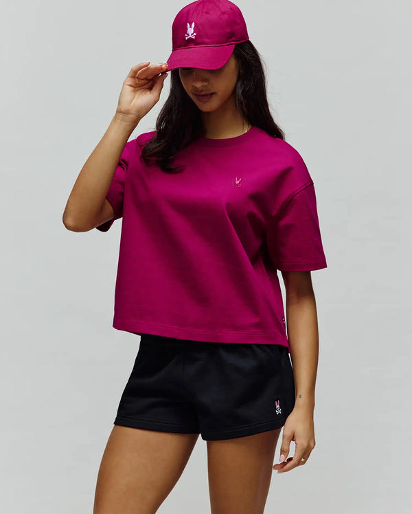 Women's Camille Oversized Fit Tee in magenta jersey with a small embroidered Psycho Bunny logo on the chest, paired with black athletic shorts featuring a pink bunny logo and a matching magenta baseball cap with a white embroidered logo.