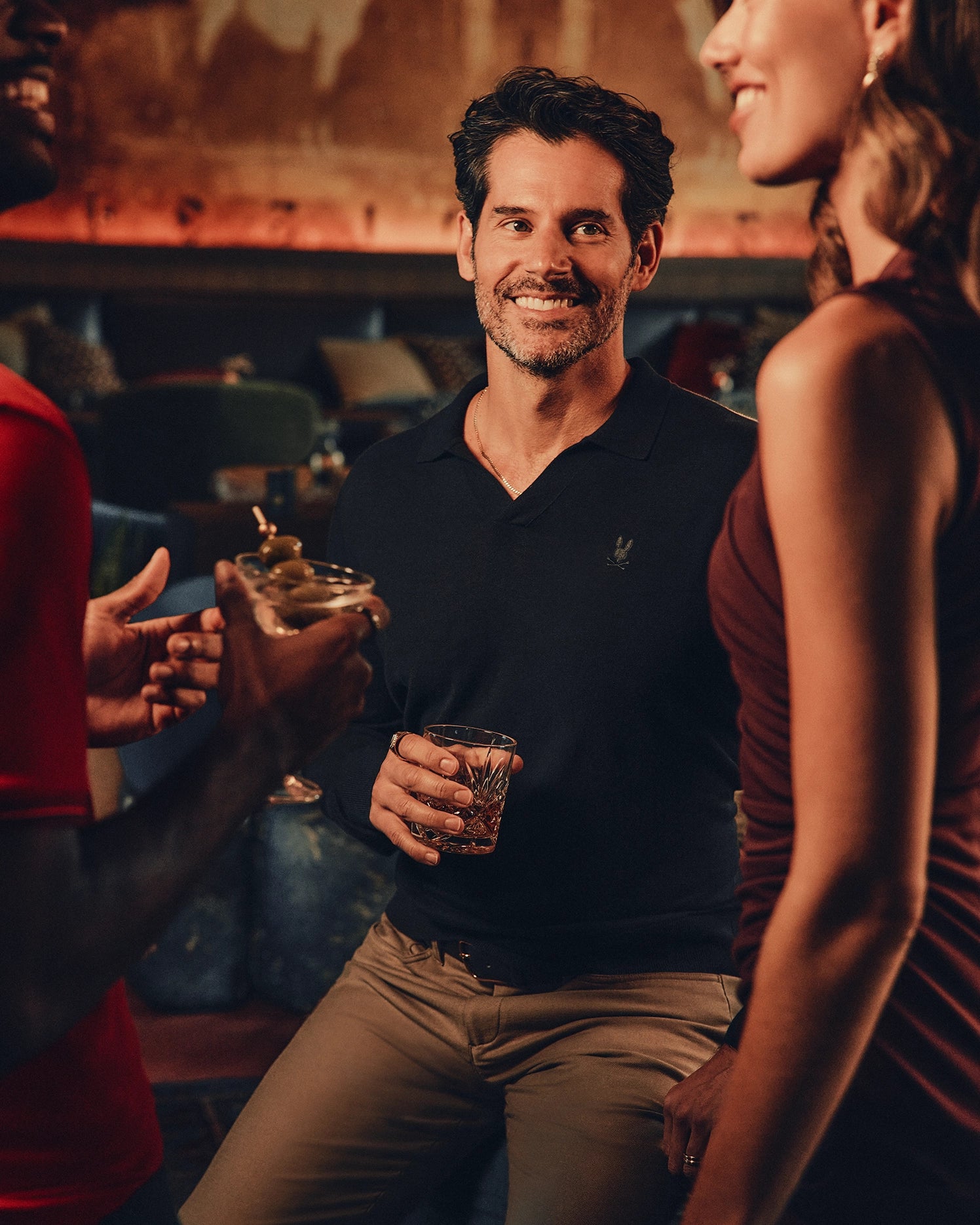 A man in a Psycho Bunny MENS JOHNNY COLLAR SWEATER - B6E258E200 and beige pants holds a drink and smiles, surrounded by two people chatting in a warmly lit bar or lounge.