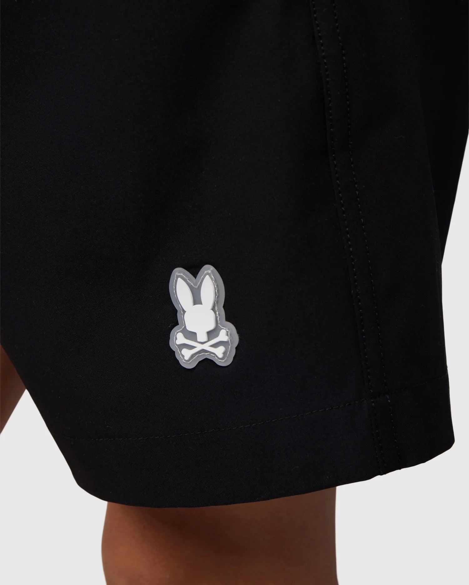 Close-up of the hem of a pair of Psycho Bunny KIDS MALTA HYDROCHROMIC SWIM TRUNK - B0W321B2SW, featuring a small bunny head logo with crossed bones underneath. The logo is white with a gray outline and stands out against the black fabric. The subtle bunny pattern offers a unique touch, while the background remains plain and unobtrusive.