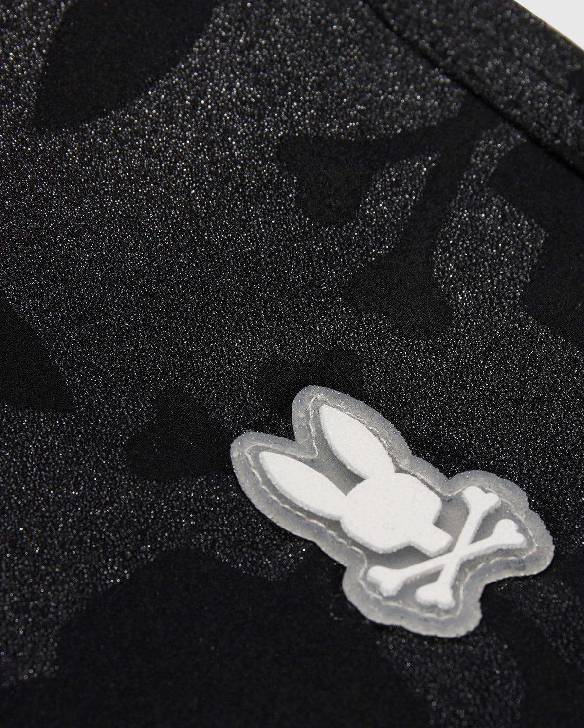 The KIDS MALTA HYDROCHROMIC SWIM TRUNK - B0W321B2SW by Psycho Bunny showcases a close-up of textured black fabric with a subtle Bunny pattern. A small, white, cartoon-style emblem displays a rabbit head with crossed bones beneath it, resembling a playful take on the skull and crossbones design—perfect for stylish swim trunks.