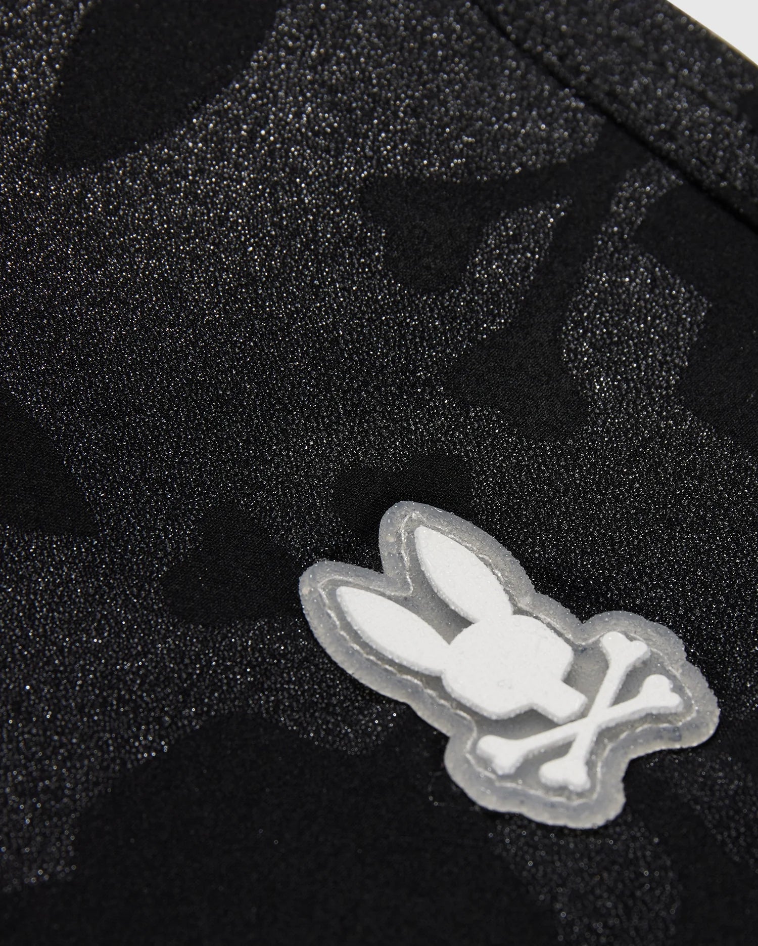 The KIDS MALTA HYDROCHROMIC SWIM TRUNK - B0W321B2SW by Psycho Bunny showcases a close-up of textured black fabric with a subtle Bunny pattern. A small, white, cartoon-style emblem displays a rabbit head with crossed bones beneath it, resembling a playful take on the skull and crossbones design—perfect for stylish swim trunks.