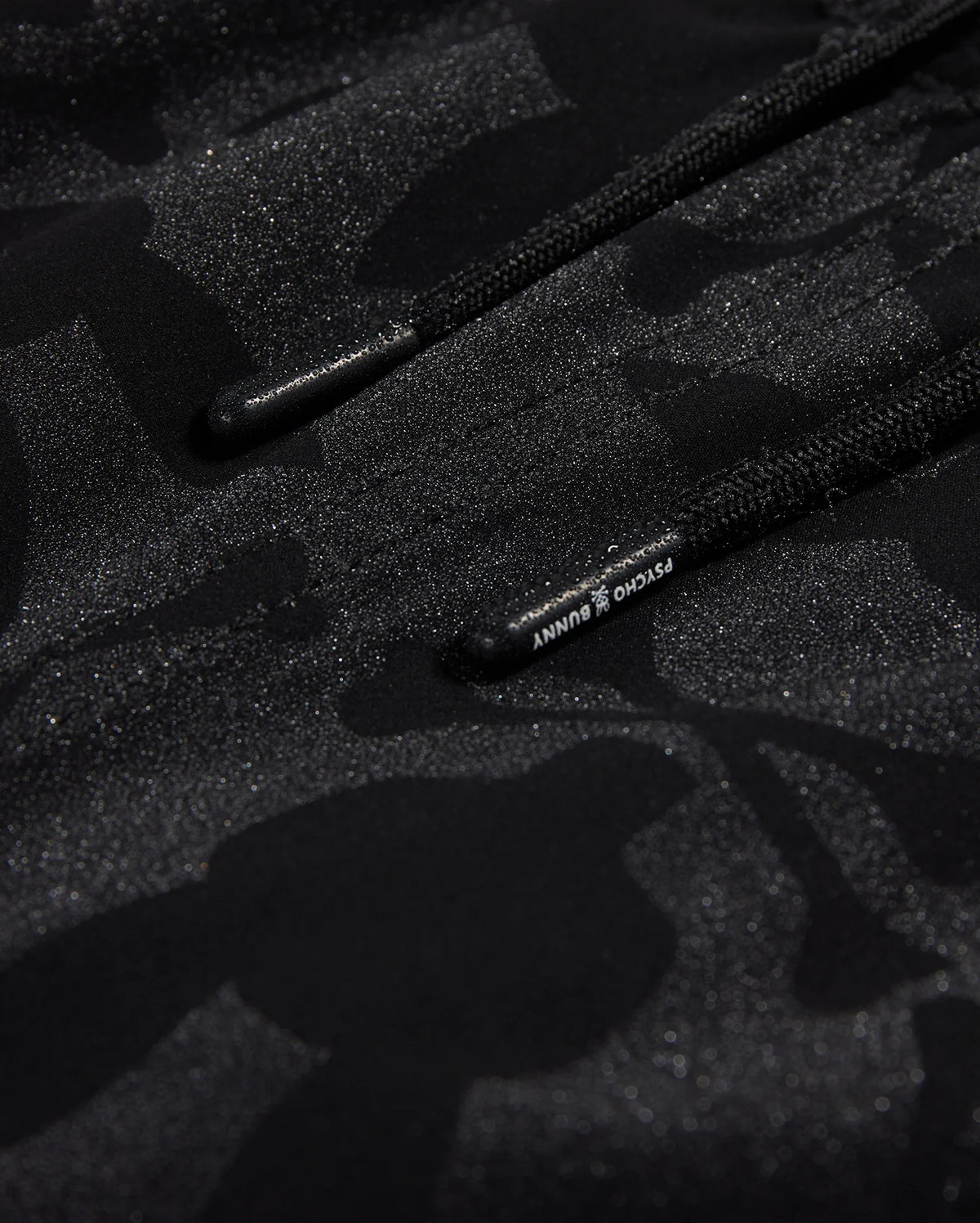 Close-up of Psycho Bunny's KIDS MALTA HYDROCHROMIC SWIM TRUNK - B0W321B2SW, showcasing a black textured fabric with a subtle camouflage pattern in shades of dark grey and black. Two parallel black cords with small text on one cord are partially visible, reminiscent of sleek swim trunks. The overall appearance is both modern and hydrochromic.
