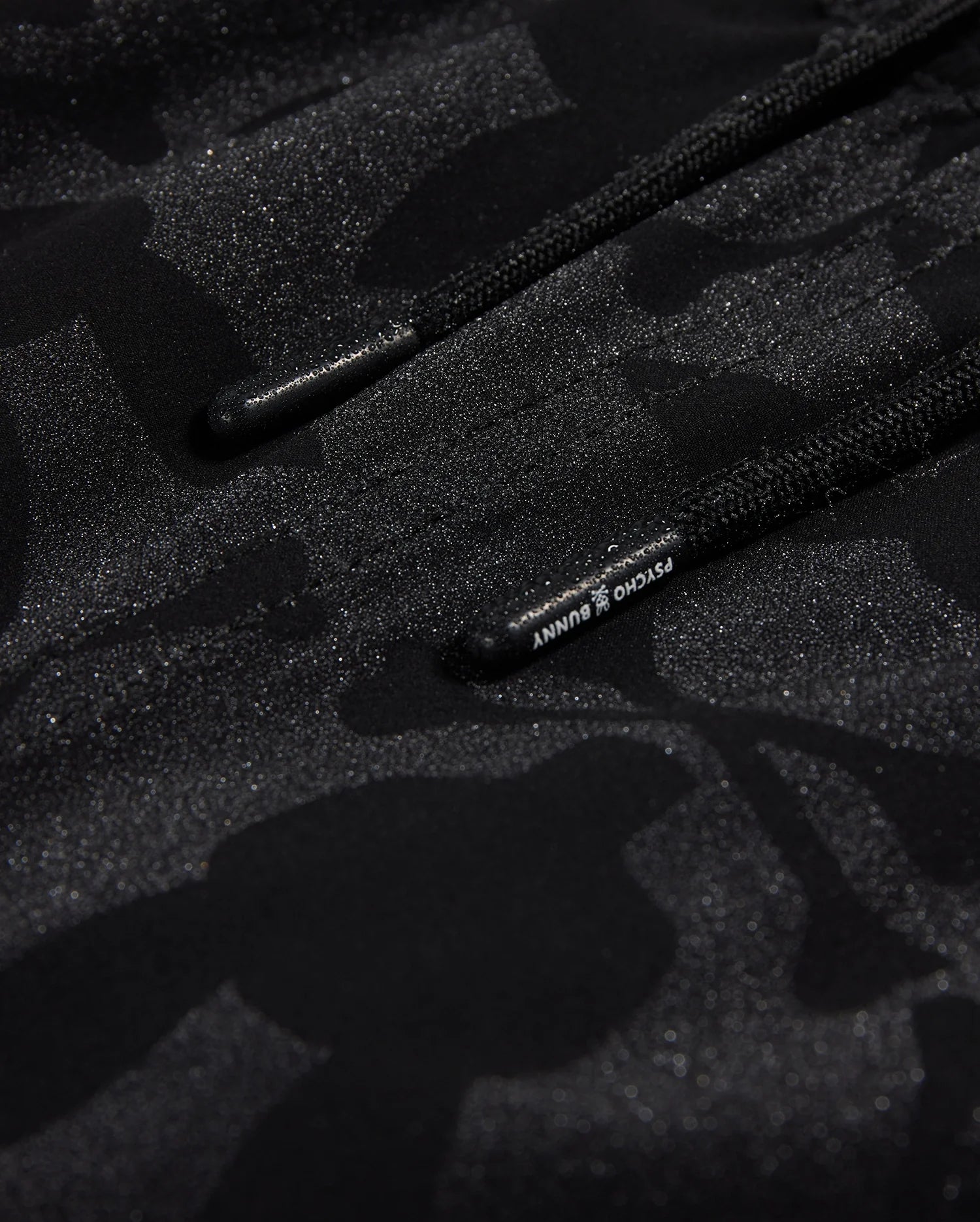 Close-up of Psycho Bunny's KIDS MALTA HYDROCHROMIC SWIM TRUNK - B0W321B2SW, showcasing a black textured fabric with a subtle camouflage pattern in shades of dark grey and black. Two parallel black cords with small text on one cord are partially visible, reminiscent of sleek swim trunks. The overall appearance is both modern and hydrochromic.