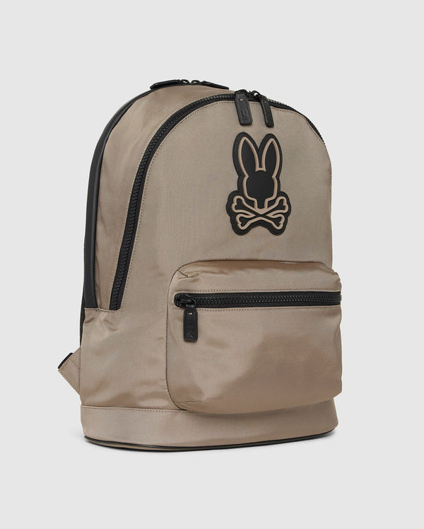 The Psycho Bunny DOME BACKPACK - B6A628Z1BP, in beige with black zippers, features a bold black bunny logo with crossbones, a padded laptop compartment, front zip pocket, and side pocket on a plain light background.