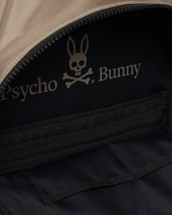 Close-up of the DOME BACKPACK - B6A628Z1BP by Psycho Bunny, featuring the official bunny skull and crossbones logo, zippered pockets, padded laptop compartment, and beige/black fabric interior.