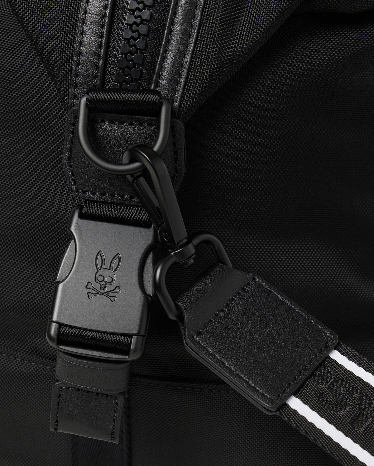 Close-up of the versatile Psycho Bunny MENS WEEKENDER BAG - B6A629Z1BG in black fabric, featuring a metal clip and buckle with a signature skull and bunny ears logo. Black leather strap with white stitching attaches to the buckle, highlighting the zipper and fabric texture.