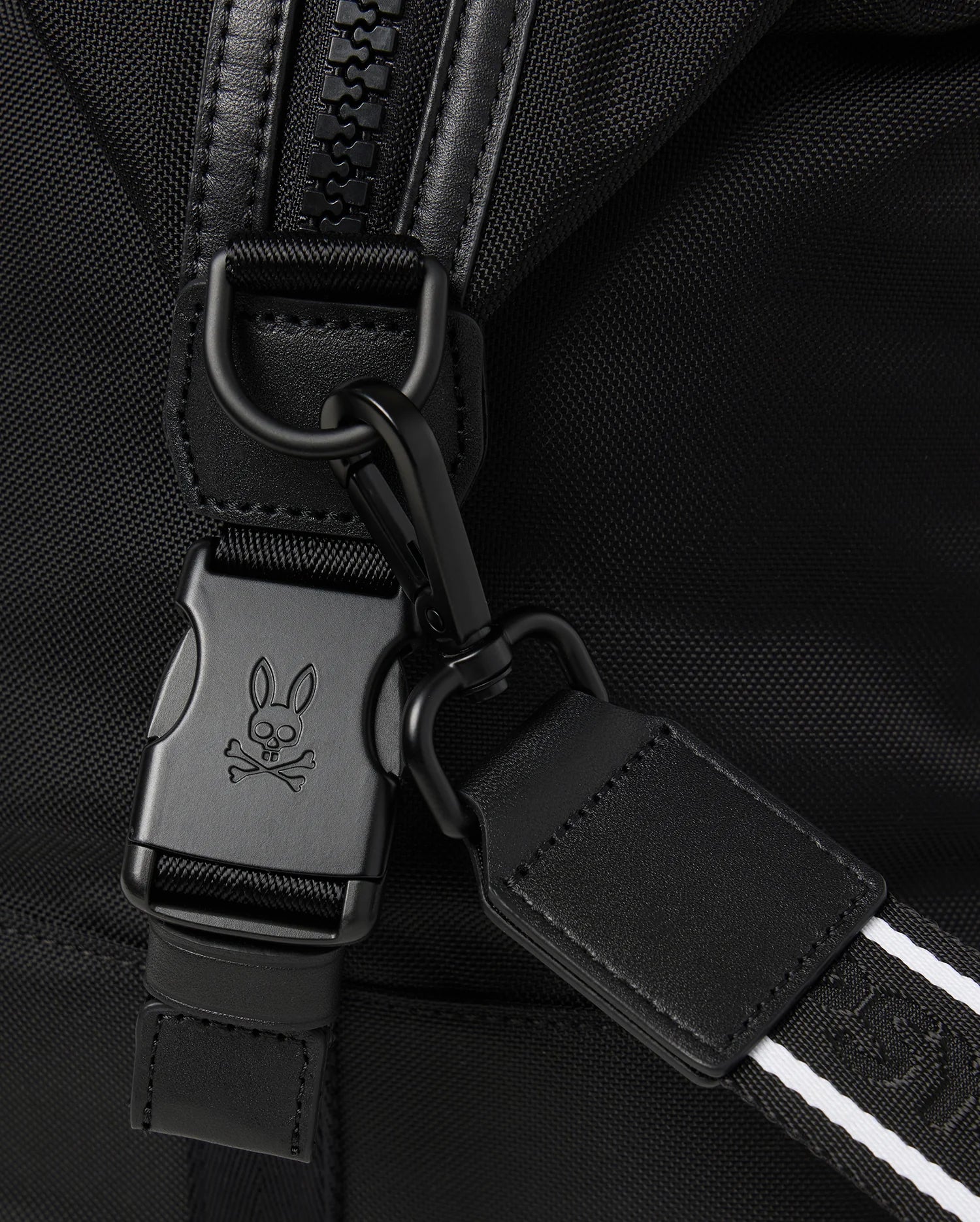 Close-up of the versatile Psycho Bunny MENS WEEKENDER BAG - B6A629Z1BG in black fabric, featuring a metal clip and buckle with a signature skull and bunny ears logo. Black leather strap with white stitching attaches to the buckle, highlighting the zipper and fabric texture.
