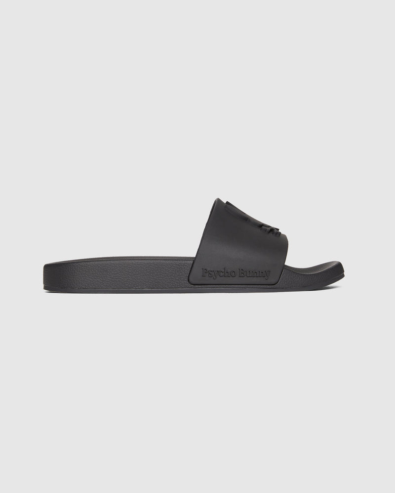 The MENS BUNNY LOGO SLIDES - B6A910U1FT by Psycho Bunny are black rubber pool slide sandals with a wide strap featuring embossed Psycho Bunny logos. The flat, textured sole provides grip and comfort, while the molded footbed ensures a perfect fit. The words "Psycho Bunny" are prominently embossed on the side of the strap against a plain white background.