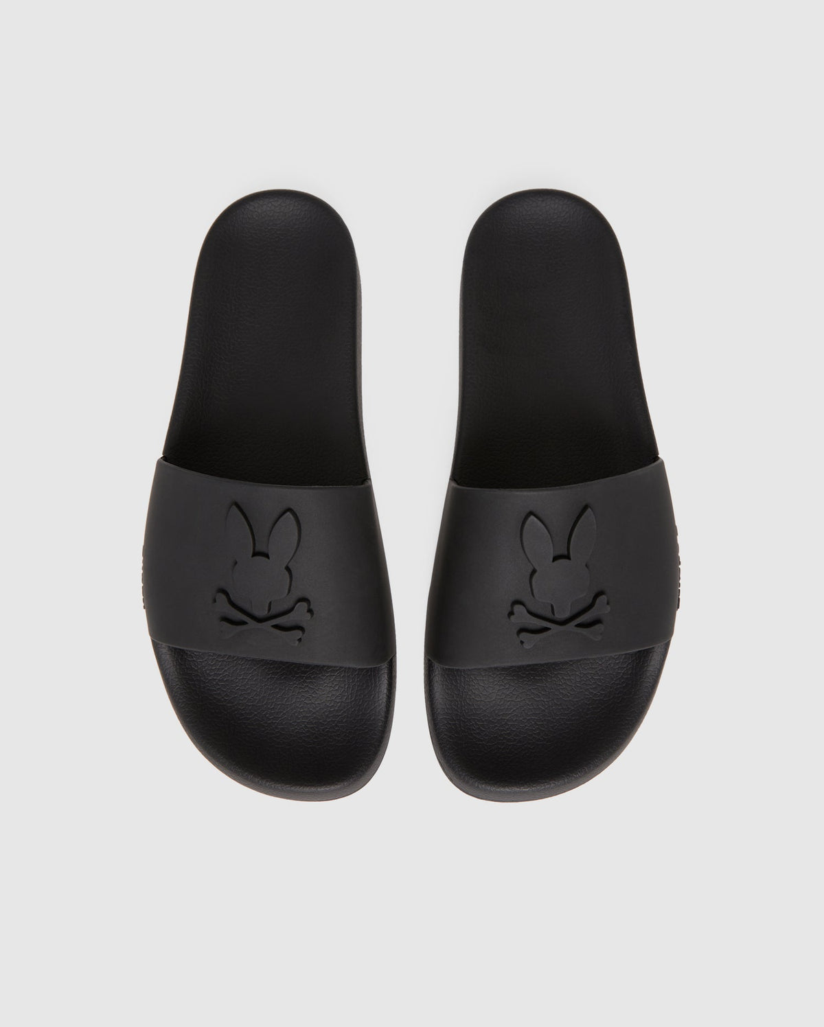 Presenting the Psycho Bunny MENS BUNNY LOGO SLIDES - B6A910U1FT: A pair of black slip-on rubber pool slides boasting a minimalist design. The top straps feature embossed logos with a stylized bunny face and crossed bones beneath. These sandals offer a textured insole and molded footbed for enhanced comfort.