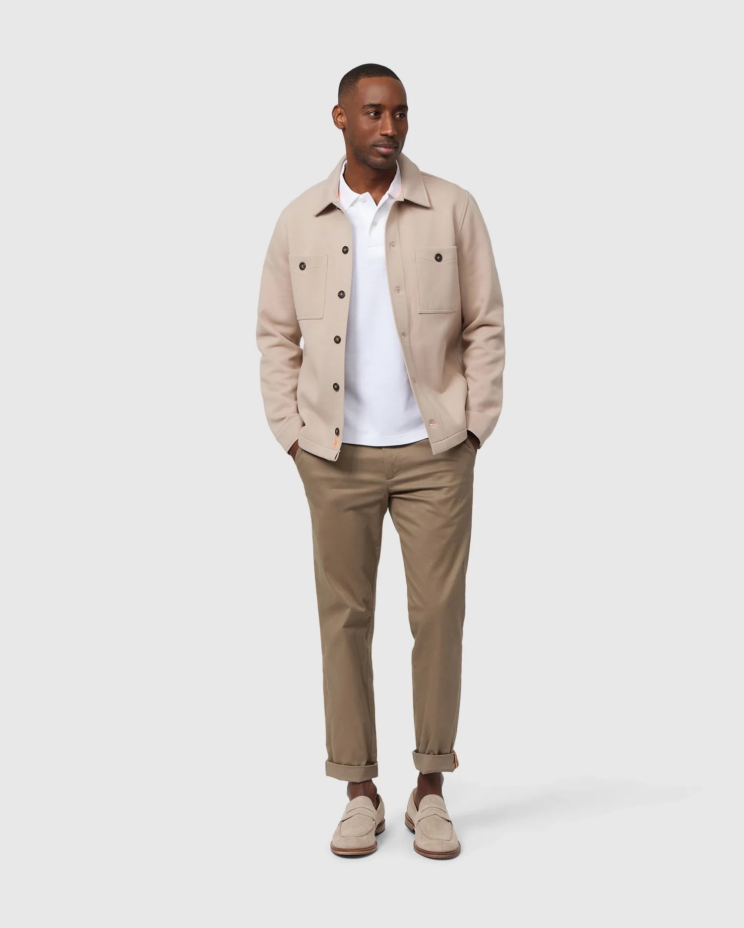 A man stands against a plain background, showcasing an elevated style with the Psycho Bunny Men's Bedford Shirt Jacket in beige (product code: B6J290B200) over a white shirt, paired with khaki pants. He has a relaxed posture with his hands in his pockets and completes the look with light-colored shoes and matching socks for an exceptional on-body feel.